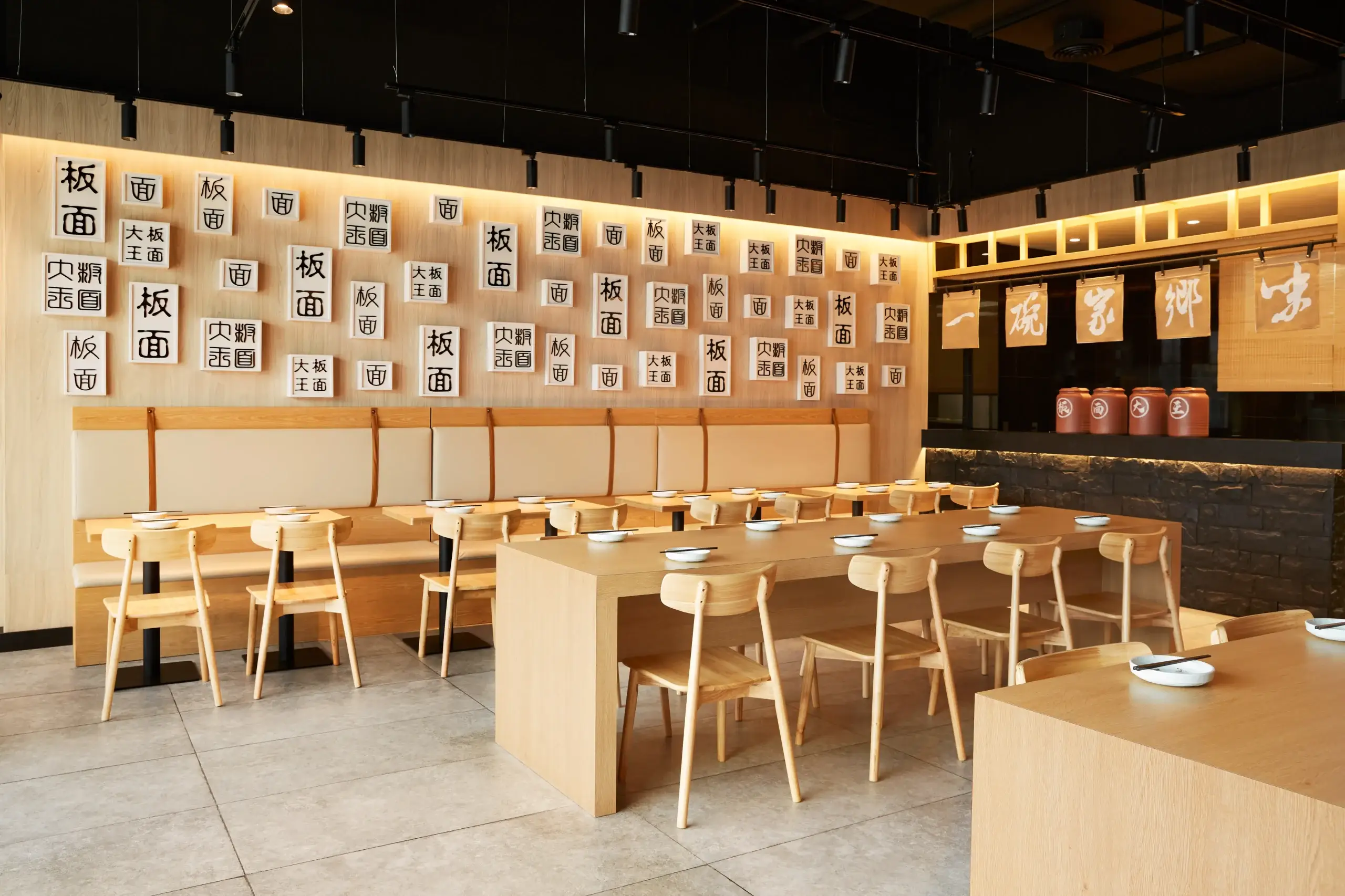 A warm, wooden Japanese restaurant interior with long communal tables and stools. The wall features numerous white panels with black kanji characters, and a counter area with jars and hanging banners in the background.