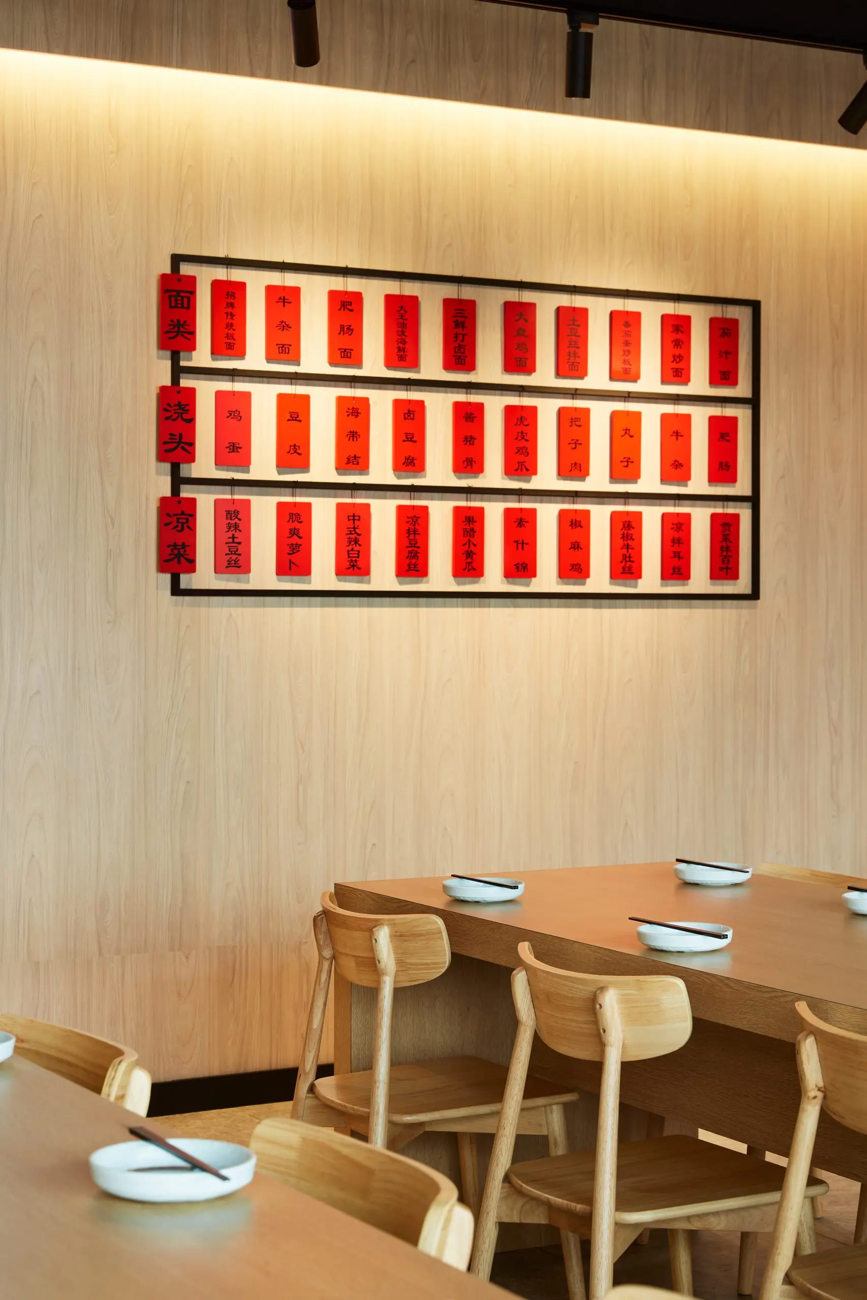 Interior of a light wood dining area with a black framed board displaying multiple red vertical scrolls with Chinese characters, mounted on a beige wall above wooden tables and chairs set with white plates and black chopsticks.