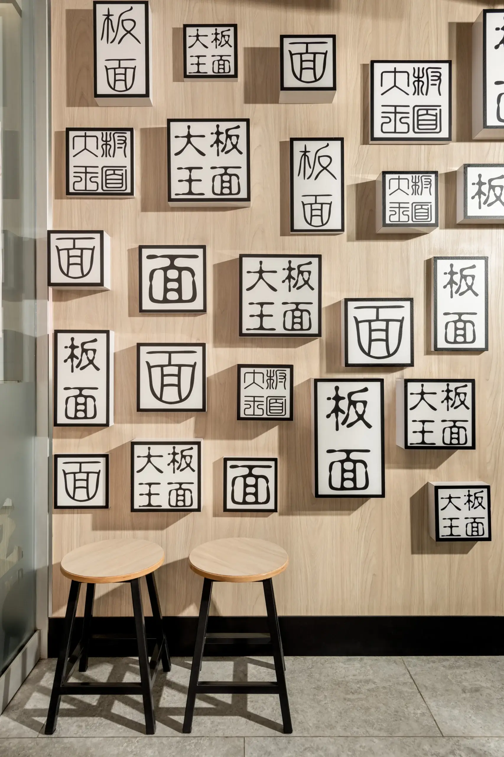 A wooden wall covered with many framed square signs bearing bold black Chinese characters of varying sizes and arrangements. Two wooden stools sit in front on a tiled floor.