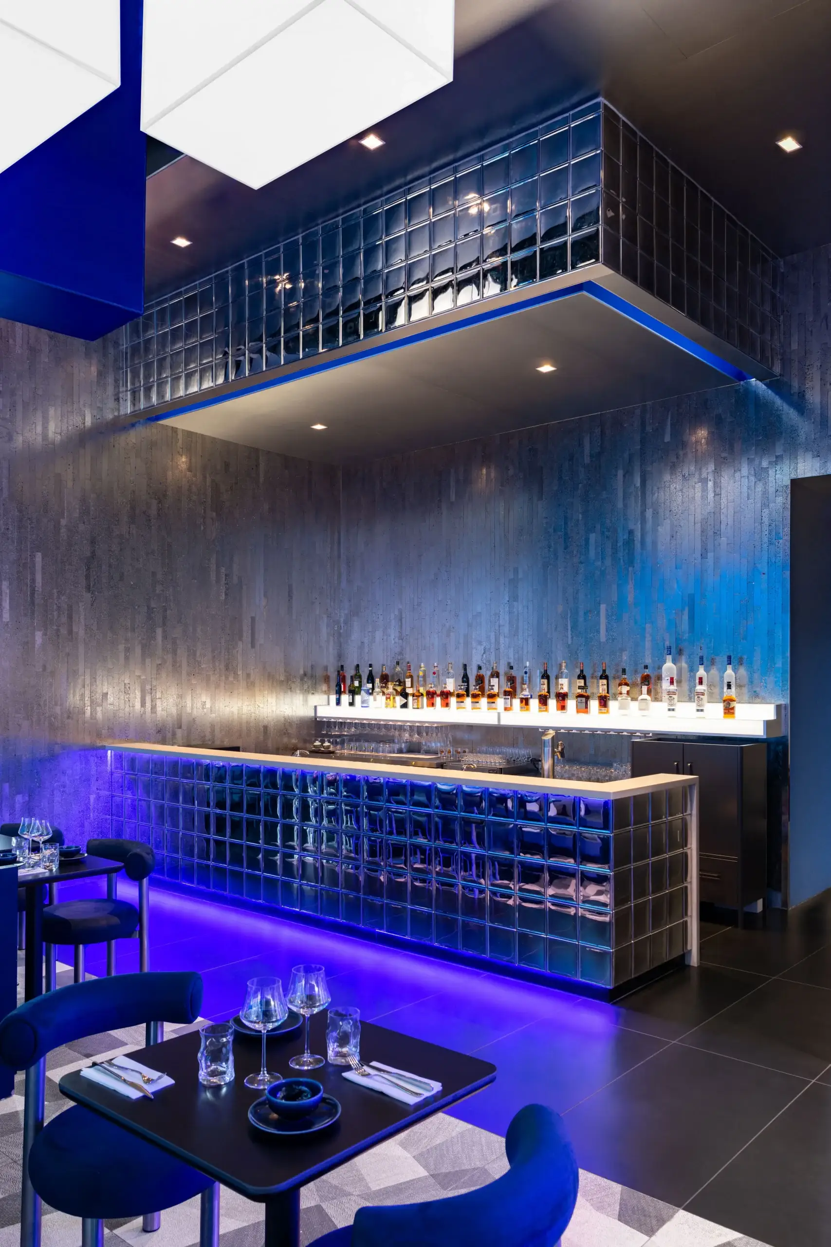 Stylish bar with a long glass-tiled counter, blue accent lighting, and a backlit shelf of assorted liquor bottles. Modern seating and dark tiled walls create a sophisticated ambiance.