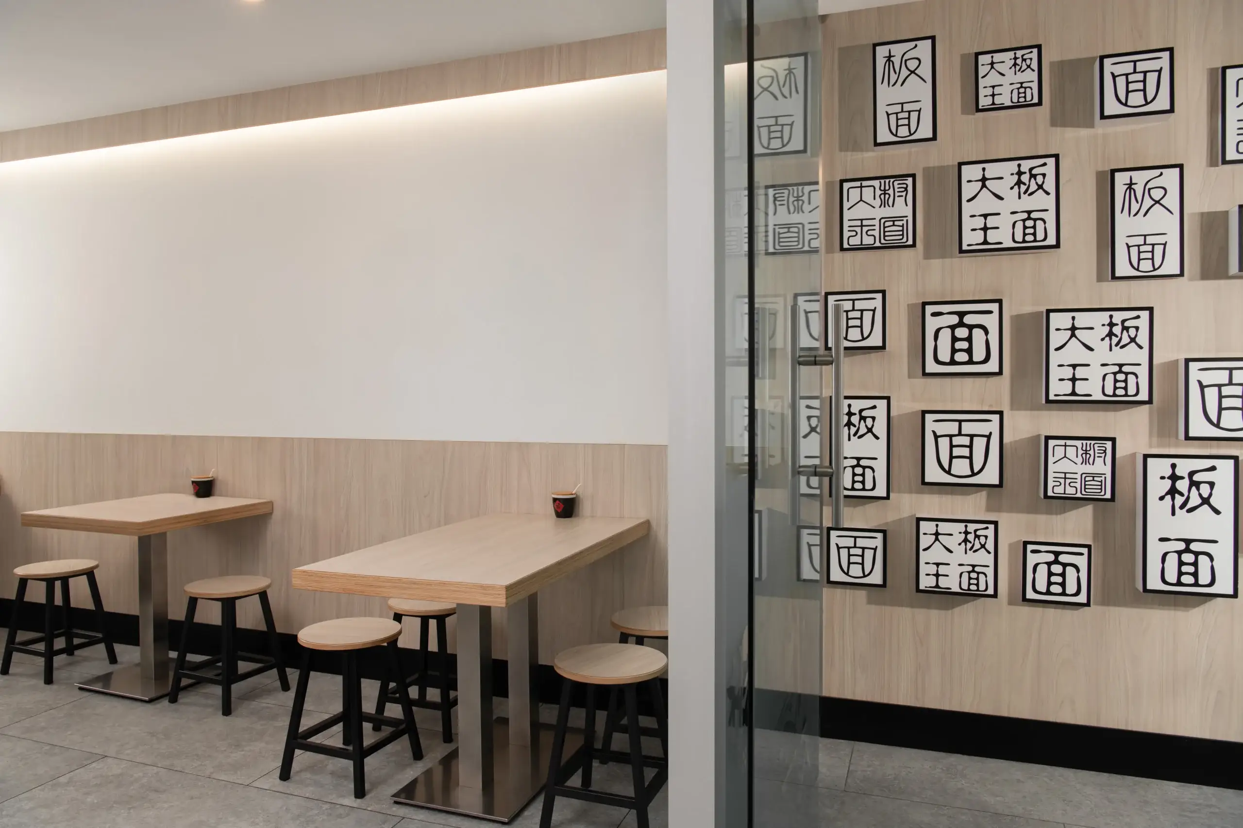A clean, modern restaurant interior featuring light wood tables and matching stools arranged along a white wall with warm ambient lighting. A glass divider on the right displays framed black-and-white calligraphic characters on a wooden panel.